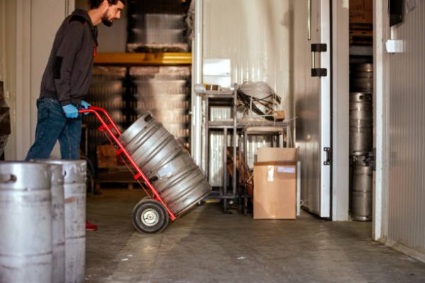 Young man working in brewery warehouse, pushing aluminum barrels on cart